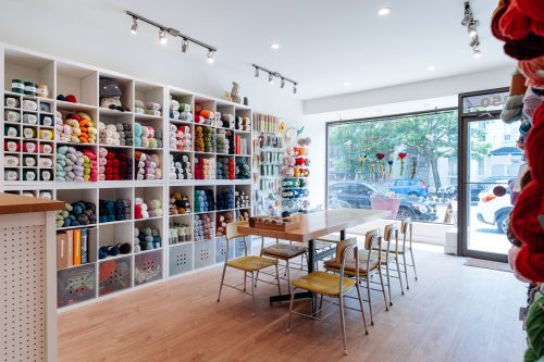 Interior view of a shop with colourful wool and knitting supplies, a long work table and chairs looking towards a large window and the door to the shop.