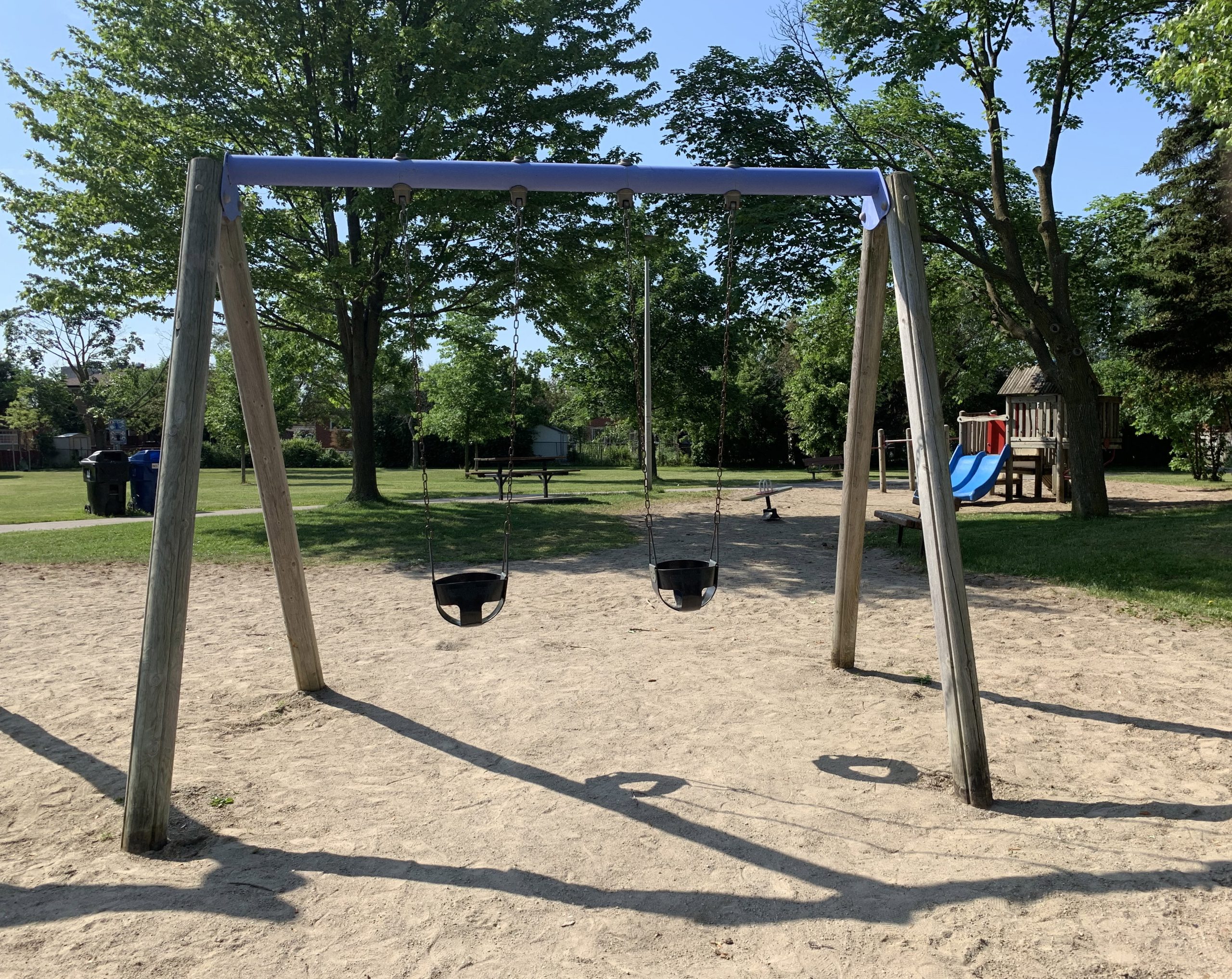 Clydesdale Park Playground before construction looking south. A wood swing set is in the foreground surrounded by sand surfacing and trees.