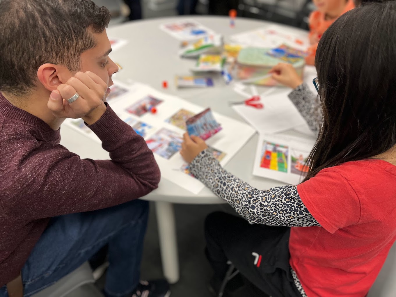 A photo of children sitting around a white table, working on a photo montage that showcases their preferences and ideas for the playground. The table is covered with colorful markers and papers with drawings and writings on them.