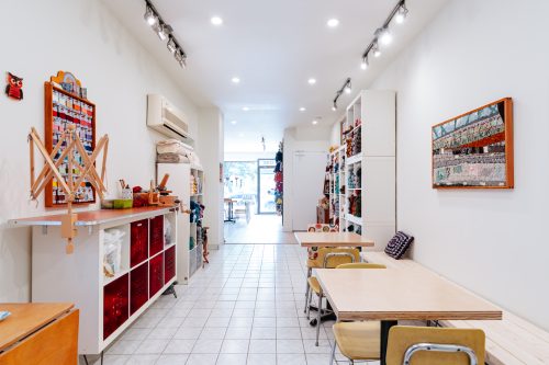 Interior view of a knitting shop from the back looking towards the front window and entrance with work tables and chairs.