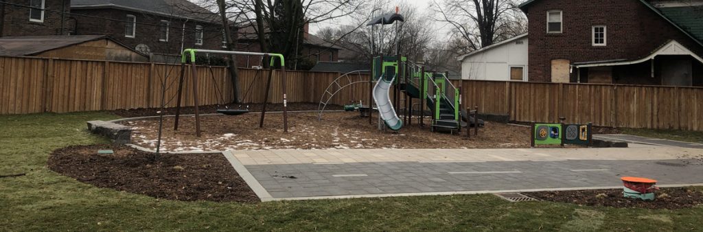 lawn. From left to right, the view of the playground includes a group swing, In front of the playground is a planting bed, park pathway and lawn.