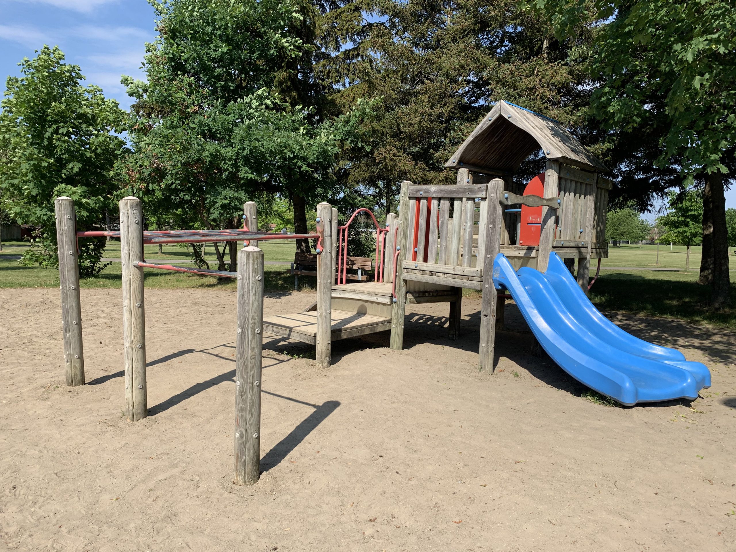 Clydesdale Park Playground before construction looking west. A wood playground is in the foreground surrounded by sand surfacing and trees.