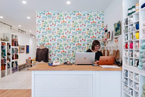 Interior view of a knitting shop with a woman working on a laptop at the front counter.