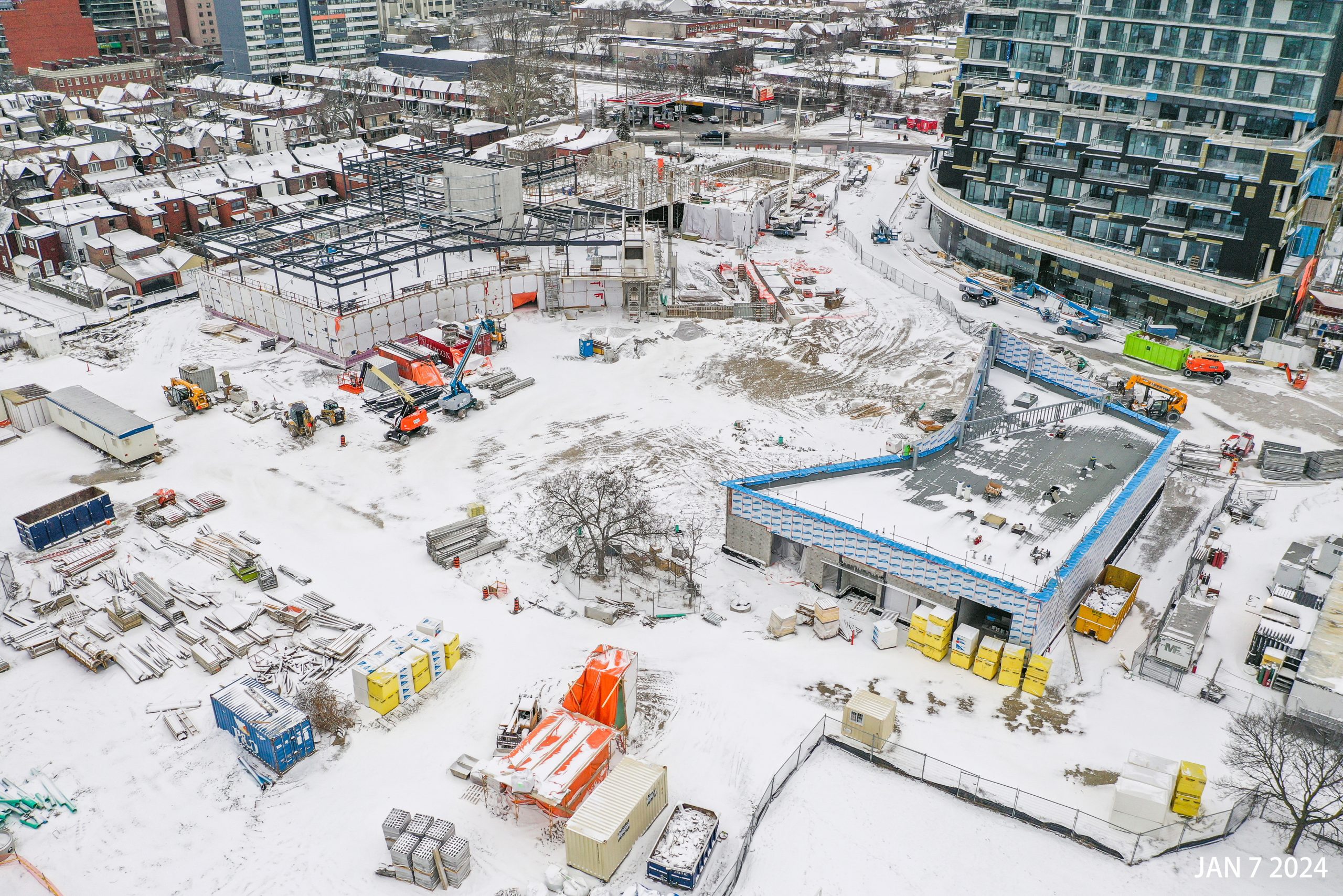 An aerial photograph of the construction site at Wallace Emersion Community Centre on January 7, 2024, which focuses on the community centre while under construction and the adjacent Fieldhouse. The community recreation structure is two-storeys with vertical beams being installed to the future third floor. The ground-level of the Fieldhouse is enclosed with an unfinished roof. The entire area is covered in a think layer of snow. 