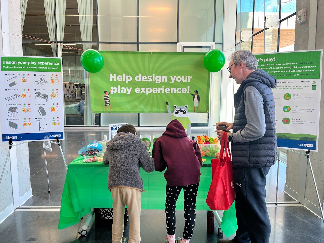 A photo of a green table with different materials and information for designing a play experience, surrounded by four green balloons. Two children and an adult are interacting with the table, while a large banner and two boards on the wall behind them invite others to participate in the activities.