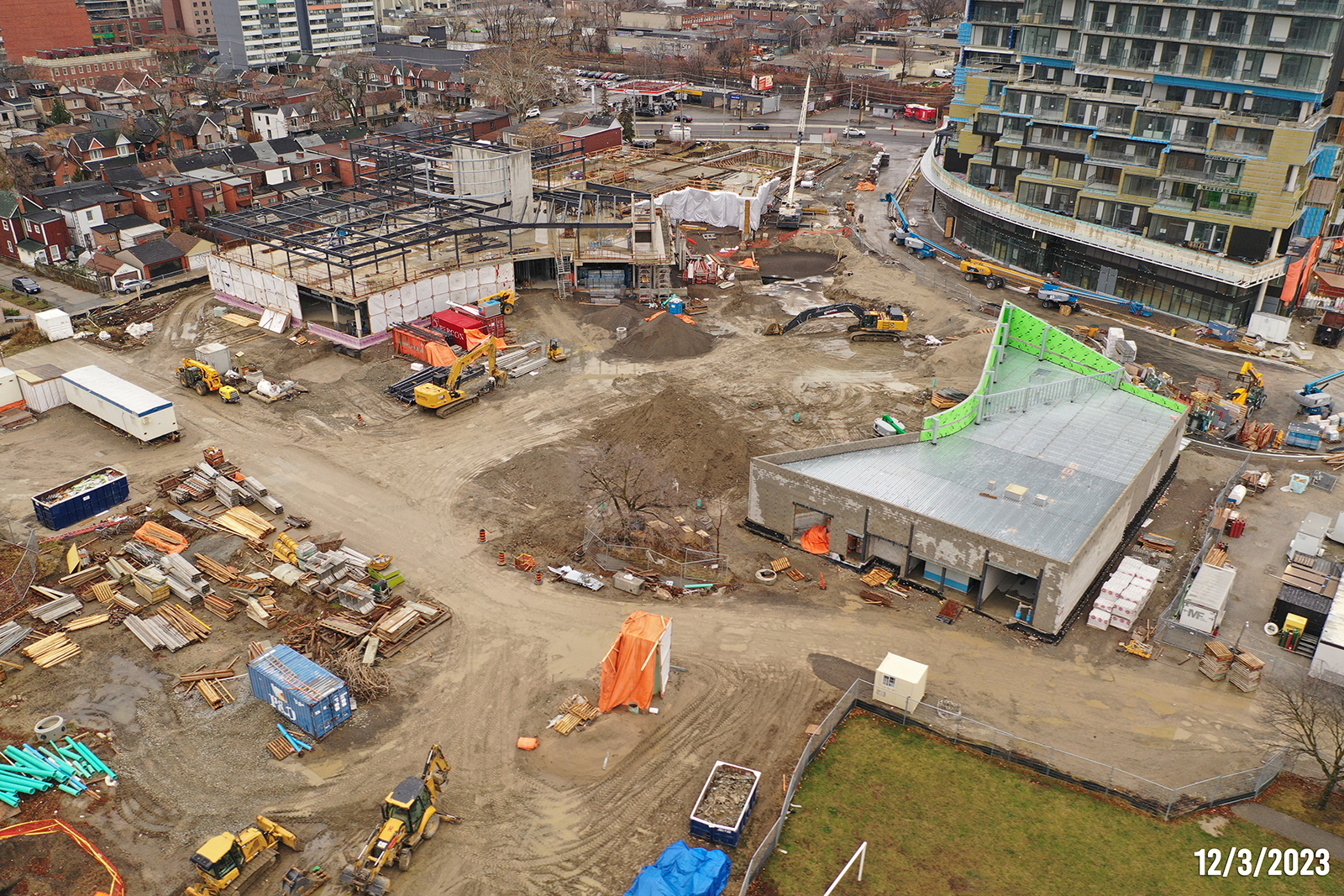 An aerial photograph of the construction site at Wallace Emersion Community Centre on December 3, 2023, which focuses on the community centre while under construction and the adjacent Fieldhouse. The community recreation structure is two-storeys with vertical beams being installed to the future third floor. The ground-level of the Fieldhouse is enclosed with an unfinished roof. 