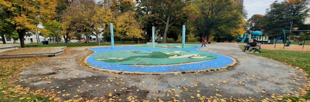 Dentonia Park splash pad in the fall which shows a circular water play area on blue and green coloured concrete with three tall water spray poles. The splash pad area is surrounded by trees and next to the playground.