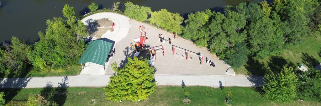 A high angle shot of Terraview Park with various features, such as a pond, a playground, a splash pad, a walking path, and a shade structure. People are seen in different activities, such as swinging, sliding, sitting, and walking.