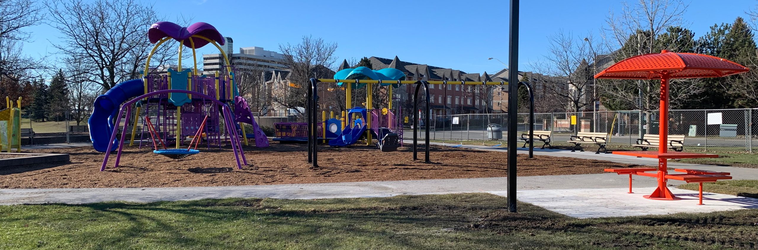 Image shows the new playground which is blue, purple, and yellow. From left to right, there is a basket swing, large senior play structure, swingset, and accessible junior structure with ramp, and an orange umbrella table.
