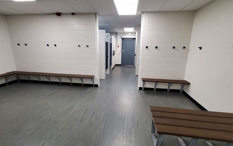 A photo of the shared pool and arena change rooms which shows grey floors, white walls with wood benches long them and the door to the pool deck in the distance.