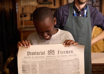 : Boy smiling and looking down at the newspaper print he is holding up. The newspaper is titled Provincial Freeman.