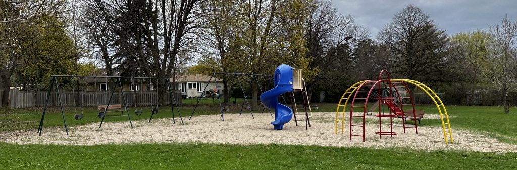Playground equipment, including two swing sets, a standalone twisting slide, and a metal spider climber on a sandy play surface as Caswell Park.