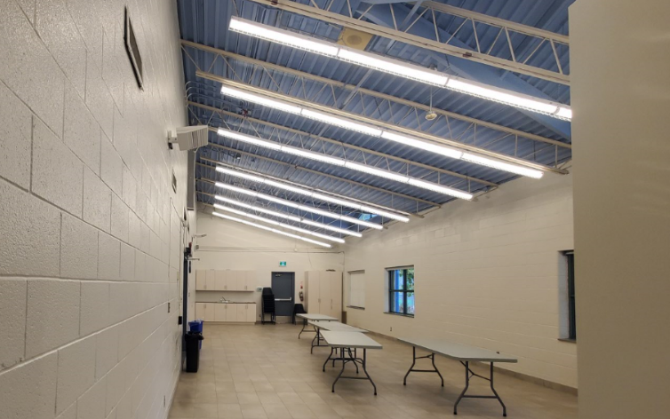 A photo of the multipurpose room in Goulding Community Centre showing a mostly empty room with four long collapsible tables in the foreground, and an exit with a blue door next to a counter with a sink and cabinets in the background.