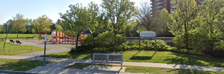 Image shows Rosebank Park before construction. There is a red and yellow playground with sand surfacing in the background, and a bench and park sign in the foreground.