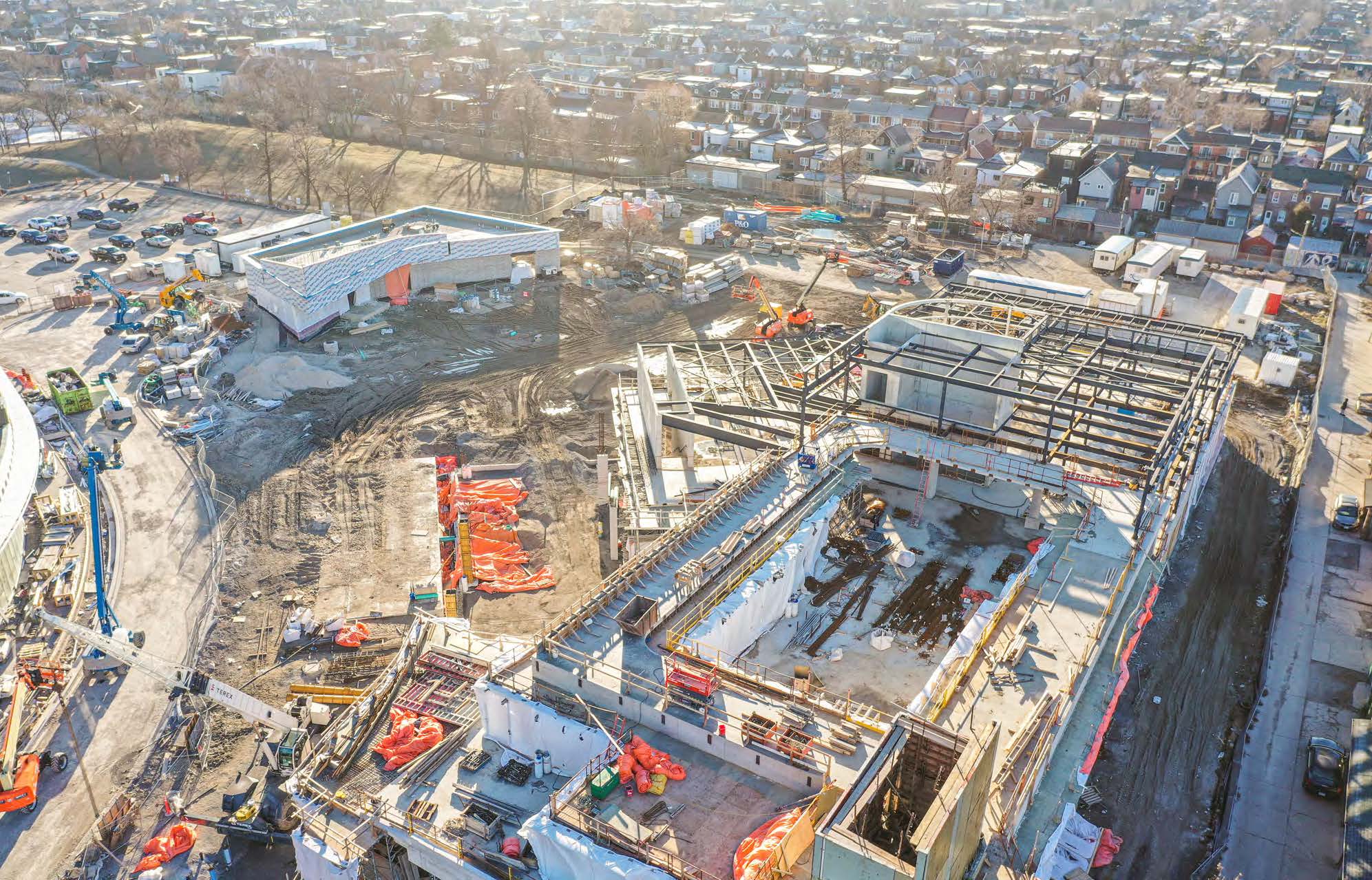 An aerial photograph of the construction site at Wallace Emersion Community Centre on February 3, 2024, which focuses on the community centre while under construction in the foreground and the adjacent Fieldhouse in the background. The community recreation structure is two-storeys partially enclosed with walls. The ground-level of the Fieldhouse is enclosed with an unfinished roof. 