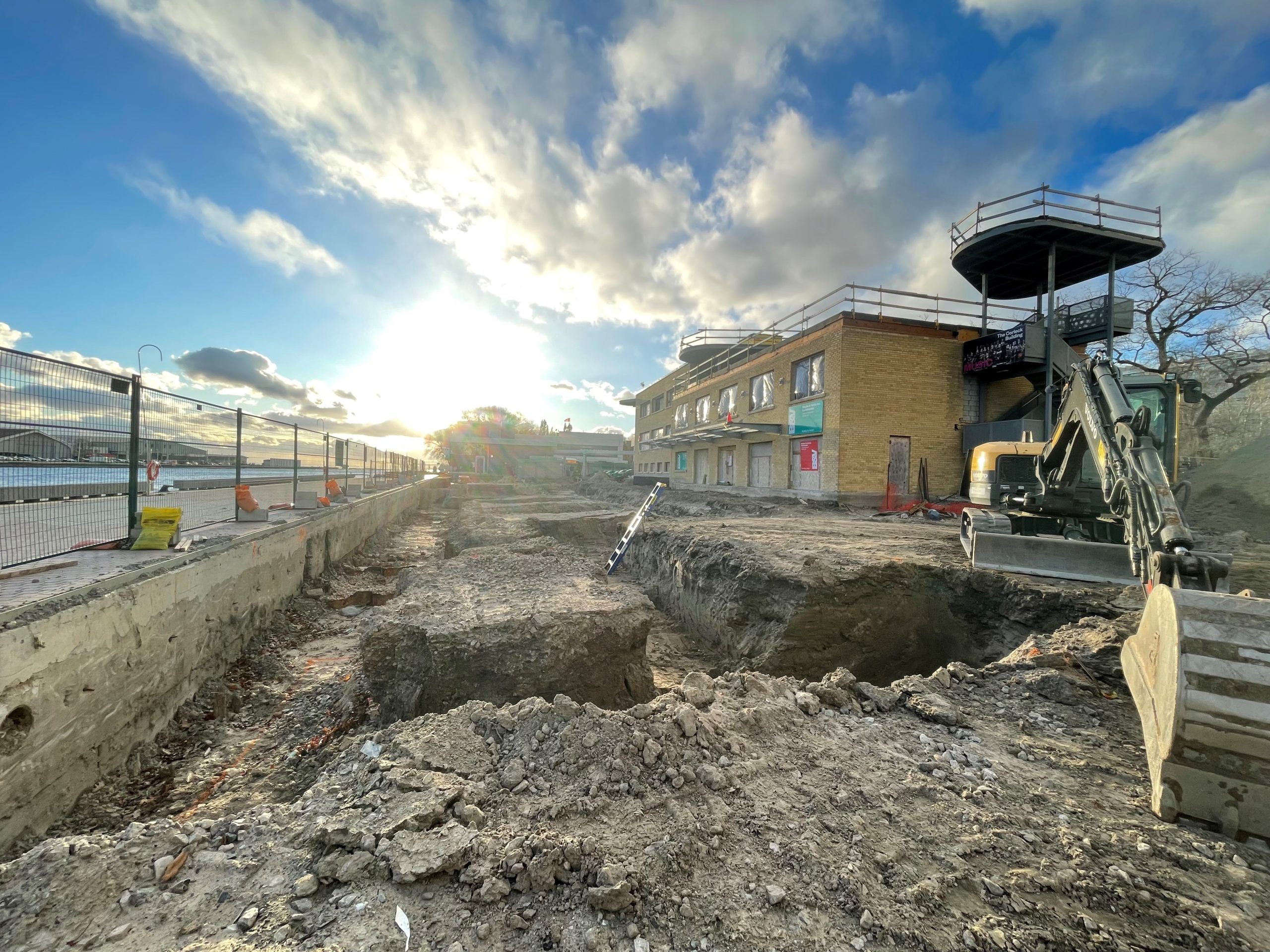 The construction site along the edge of Lake Ontario showing excavated dirt and fencing surrounding the construction area. The photo is taken facing the sun at sunrise. 