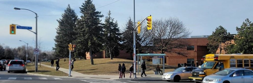 A daytime photo of the intersection of Tapscott Road and Washburn Way which includes a TTC bus stop and a school beyond the intersection. Several youth are crossing the street and walking towards the high school and many vehicles including a school bus are waiting at the red signal.
