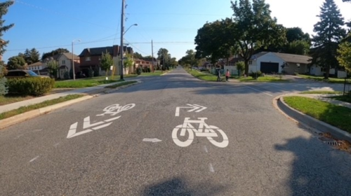 An example of on-street shared lane markings in a similar neighbourhood. The pavement markings consist of white paint showing a bicycle and directional arrows in the middle of the road. showing a 