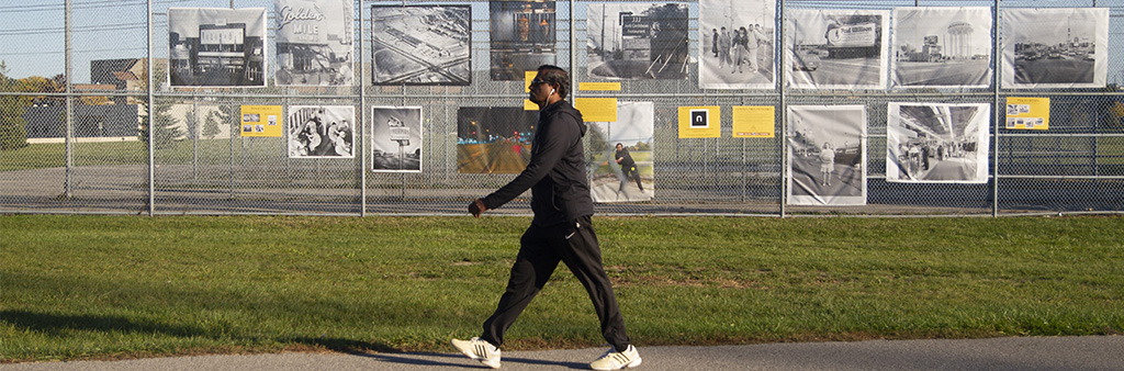 Man walks on park path in front of outdoor photography exhibition installed on chain-link fence.