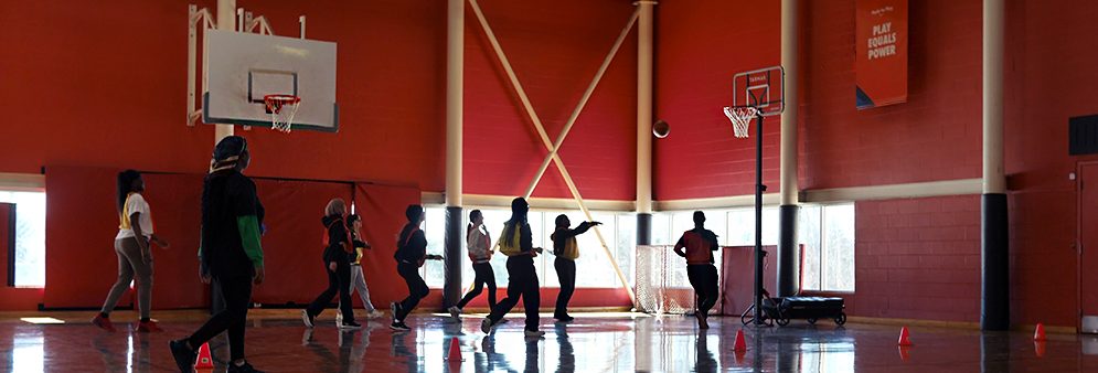 A group of girls play basketball in a gym at Ellesmere Community Centre. A banner hanging from the ceiling reads “Play Equals Power”.