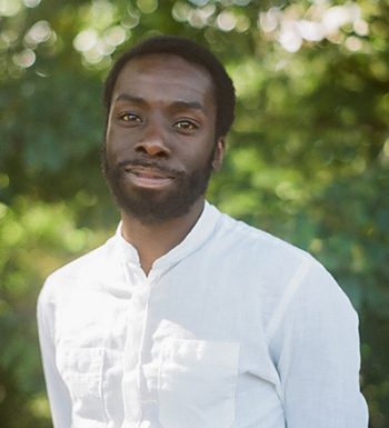 Author Desmond Cole in a white shirt in front of sun-dappled green leaves