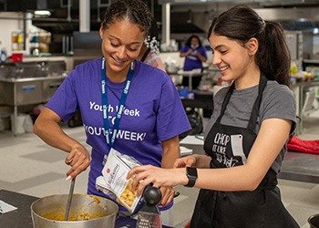 Two young people standing in front of a counter in a kitchen cooking food in a pot, wearing a volunteer T-shirt and apron.