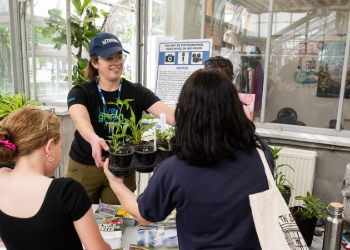 A person smiling as they hand plants in a nursery container to another person with books on the table between them and plants in the background near a glass office.