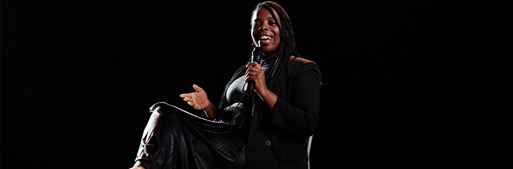 A black woman on stage holding a microphone and sitting in a chair with notes in her lap.
