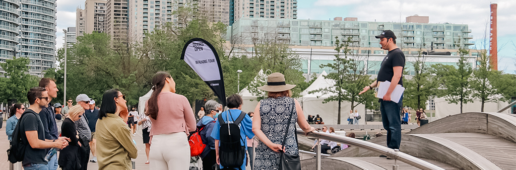 Tour group listens to guide at wavedeck on Toronto waterfront with towers in the background