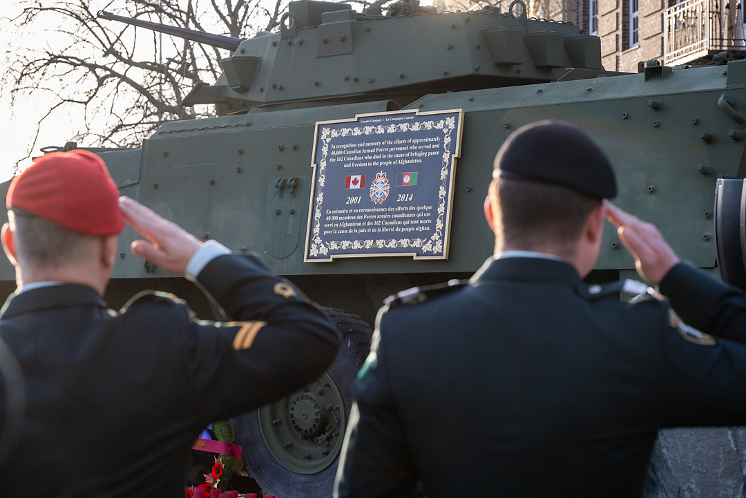 The backs of two male soldiers salute in front of the light armoured vehicle monument and plaque