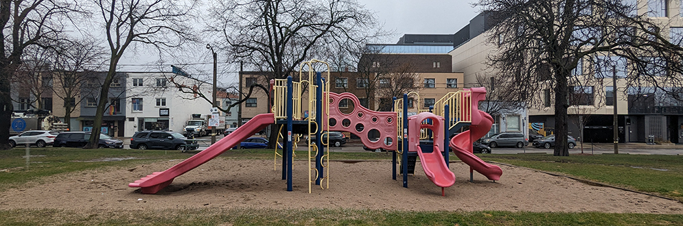 Allan A. Lamport Stadium Park Playground showing a red, yellow and blue play structure with three slides on top of sand. River Street and the business along the street are in the background.
