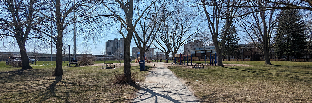 Northeast corner of Flemingdon Park taken from the pathway leading to the playground area in the background. Grass and trees are on either side of the pathway.