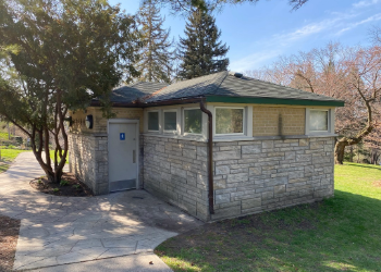 Exterior of a public washroom with stone walls near trees and greenery in a park.