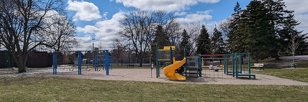 Bridlewood Park Playground showing a playground structure next to a swing set on top of sand. Grass surrounds the playground area.