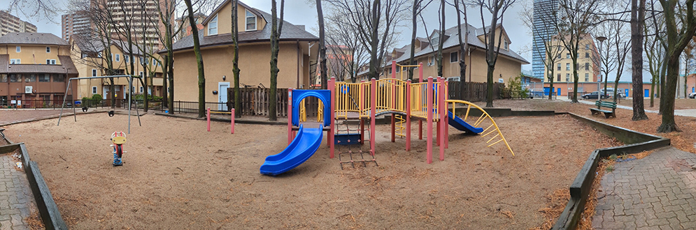 Oak Street Park Playground showing a small yellow, blue and red play structure with a small blue slide on top of sand. A spring toy and swing set are in the background.
