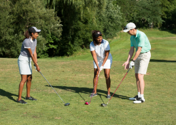 Three people holding golf clubs and standing on a grassy golf range in the address position before they take their swing.
