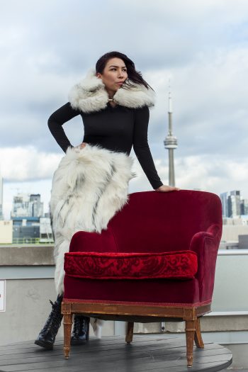 Woman wearing furs stands beside a red upholstered armchair with view of Toronto skyline in background