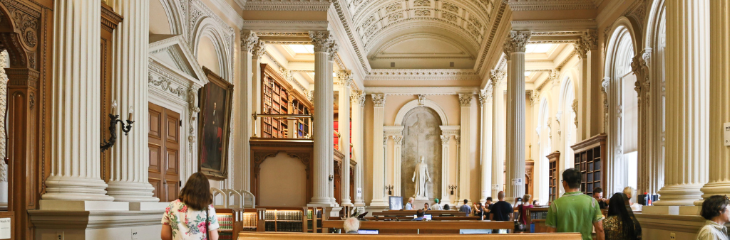 People walking through an ornate neo-classical room with Corinthian columns, vaulted ceiling and a niche with a sculpture