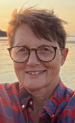 Author/editor Jane Farrow wearing a blue and red check shirt with a beach sunset behind her