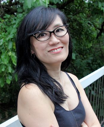 Author Kerri Sakamoto in black glasses and black sleeveless dress seated in front of green foliage