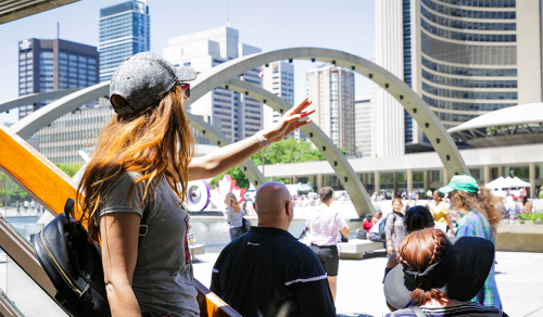 Person in foreground reaches arm towards City Hall beside group of people. Nathan Phillips Square arches and City Hall and visible in background.