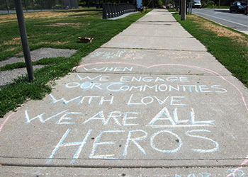 Blue and pink chalk on a stretch of sidewalk with grass on either side reading when we engage our communities with love we are all heroes.