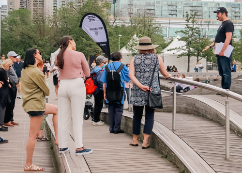 Tour group listens to guide at wavedeck on Toronto waterfront with towers in the background