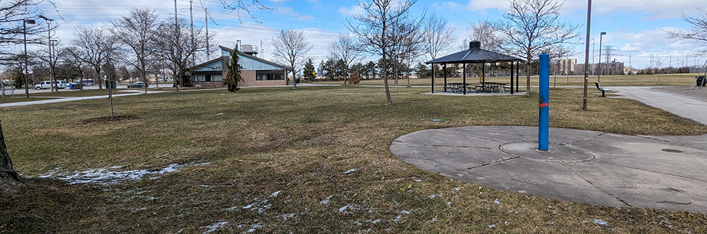 Jack Goodlad Park showing a small circular concrete pad with one spray feature in the foreground and an open lawn with trees and a shade structure in the background.