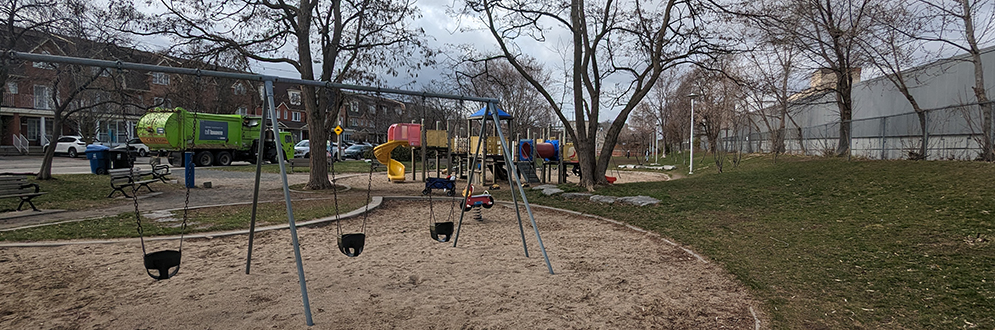 Erwin Krickhahn Park Playground showing the swing set on top of sand in the foreground and Rankin Crescent in the background.