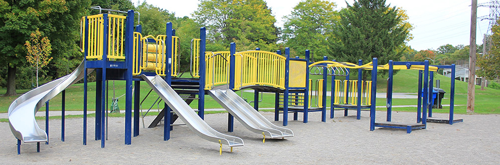 Prairie Drive Park Playground showing the large playground structure with metal slides and equipment in yellow and blue. The playground is on top of sand and surrounded by grass and mature trees.