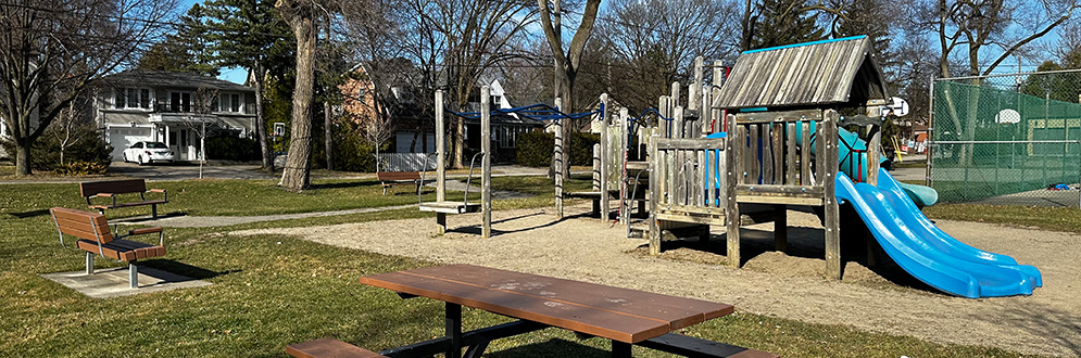 Wedgewood Park Playground with a bench in the foreground and wood play structure with a plastic blue slide in the background. Two benches face the playground.