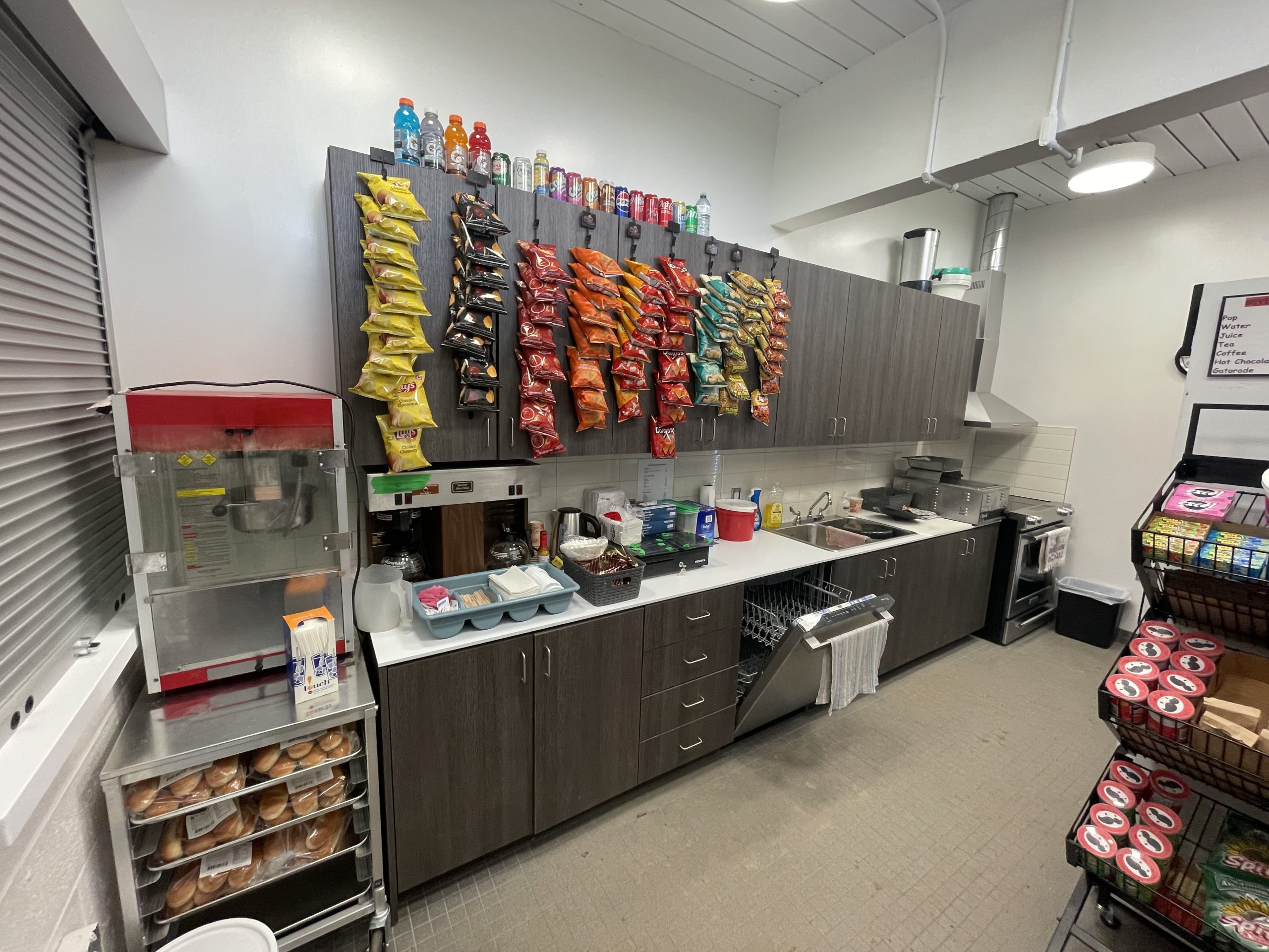 The snack bar/serving area instead the clubhouse showing a small kitchen with a dishwasher, counters, bags of chips and a popcorn machine.