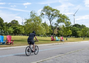 Bicyclist riding on path in front of Muskoka chairs on a grassy field and wind turbine and CN tower in background.