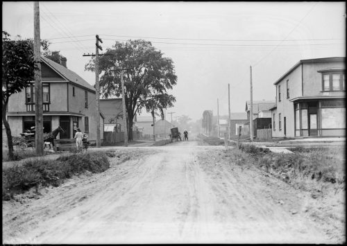 Image depicts monochrome photograph of unpaved road with two storey buildings on either side